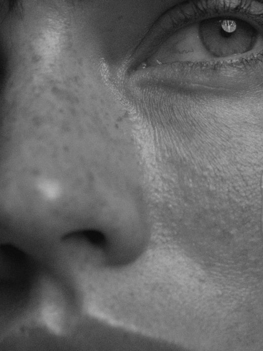 Close-up black-and-white photograph of a face in profile, showing skin texture around the eye, cheekbone, and nose in soft shadow.