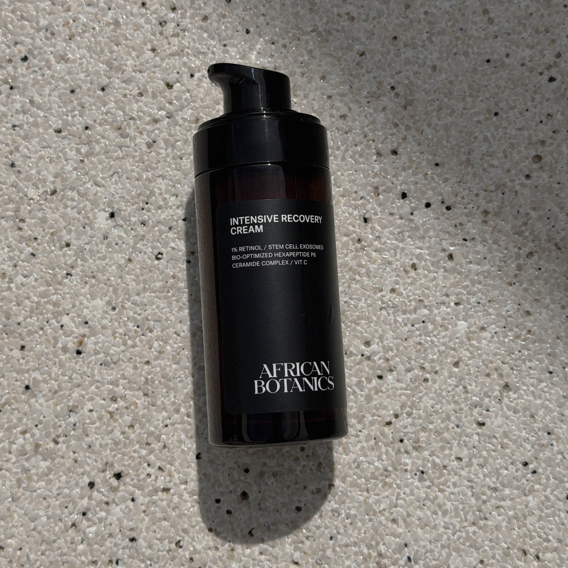 Black bottle of 'Intensive Recovery Cream' by African Botanics on a textured gray surface